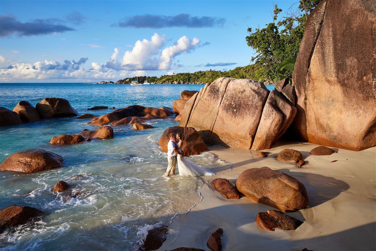 The bride and groom walk together on the beach, with majestic boulders from Seychelles islands.