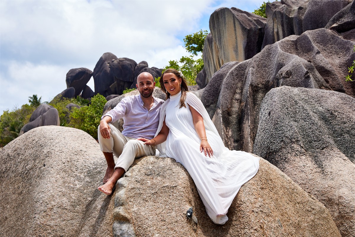 A bride and groom dressed in white are seated together on a large rock in a serene outdoor setting.