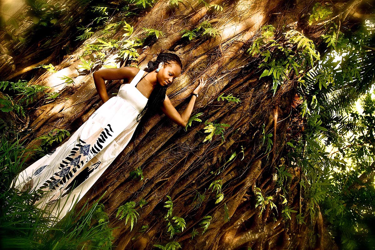 A bride wearing a white dress rests against a tree, embodying elegance and serenity in a natural setting.